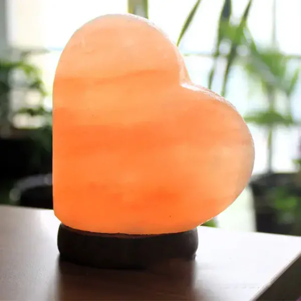 Luminária de sal rosa em forma de coração, iluminada, sobre uma superfície escura com fundo de plantas verdes.