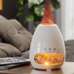 Difusor de óleo essencial em forma de gota com luz quente, contendo pedras de sal na base, colocado numa mesa de sala de estar com sofá e plantas ao fundo.