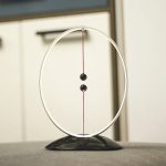 Pendulo de Newton com duas bolas pretas suspensas por fios finos dentro de um aro metálico, sobre uma superfície plana.