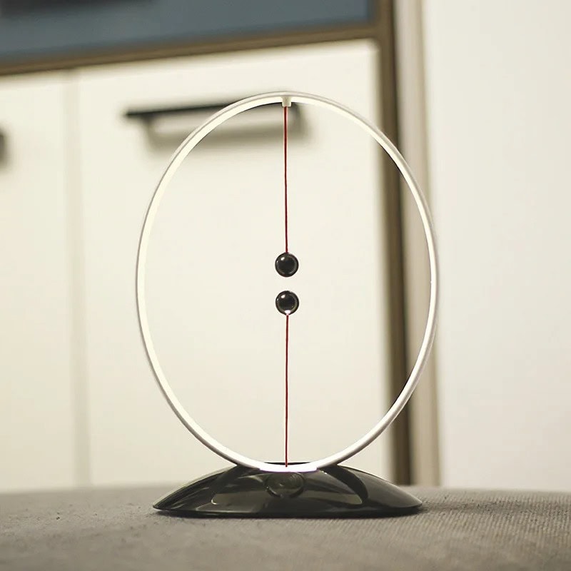 Pendulo de Newton com base decorativa Pendulo de Newton com duas bolas pretas suspensas por fios finos dentro de um aro metálico, sobre uma superfície plana.
