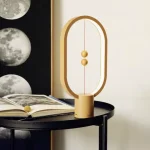 Lâmpada de decoração inspirada na ciência, em forma de órbita com esferas douradas, colocada sobre uma mesa preta com um livro aberto ao lado.