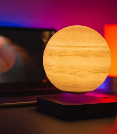 Lâmpada de planeta Júpiter com detalhes de texturas e cores, iluminada com luz quente, colocada sobre uma base preta, com fundo colorido difuso.