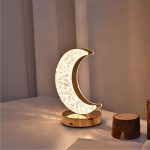 Luminária em forma de lua crescente com detalhes de estrelas, luz suave e base dourada, colocada numa mesa ao lado de um tronco de árvore decorativo.