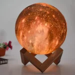 Lâmpada decorativa em forma de Lua com detalhes de crateras, iluminada com luz quente, apoiada em suporte de madeira.