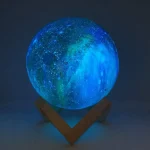 Candeeiro em forma de lua com iluminação azul e verde, apoiado por uma base de madeira, criando efeito de luz lunar.