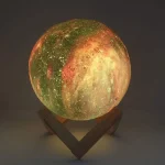 Lâmpada de forma de lua com cores verdes, vermelhas e amarelas, apoiada numa base de madeira, em ambiente escuro.