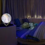 Candeeiro em forma de lua com luz suave, colocado numa mesa ao lado de uma cama com duas crianças a brincar, cortinas de fundo.