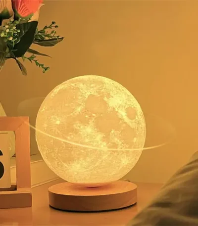 Candeeiro em forma de lua com iluminação amarelada, colocado numa base de madeira, decorando uma sala com plantas e objetos de decoração.