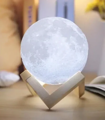Candeeiro em forma de lua com base de madeira, iluminação suave, detalhes da superfície lunar visíveis, em ambiente de sala com livros e decoração.