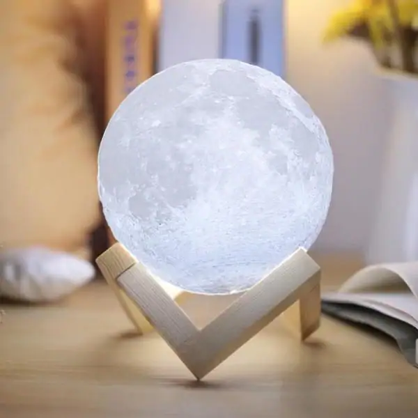 Candeeiro em forma de lua com base de madeira, iluminação suave, detalhes da superfície lunar visíveis, em ambiente de sala com livros e decoração.