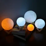 Conjunto de cinco candeeiros em forma de lua, com variações de tamanho e cor, incluindo uma luz azul, uma branca, uma laranja e uma cinza, apoiados em suportes de madeira.