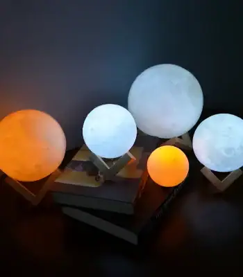 Conjunto de cinco candeeiros em forma de lua, com variações de tamanho e cor, incluindo uma luz azul, uma branca, uma laranja e uma cinza, apoiados em suportes de madeira.