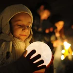 Menina com gorro e casaco segurando um candeeiro em forma de lua, iluminando seu rosto em ambiente escuro, com outras pessoas ao fundo também com luzes.