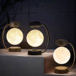 Três candeeiros lunares magnéticos com luz quente, base de metal dourado e design moderno, colocados sobre uma superfície de mármore escuro, com fundo escuro e plantas ao fundo.