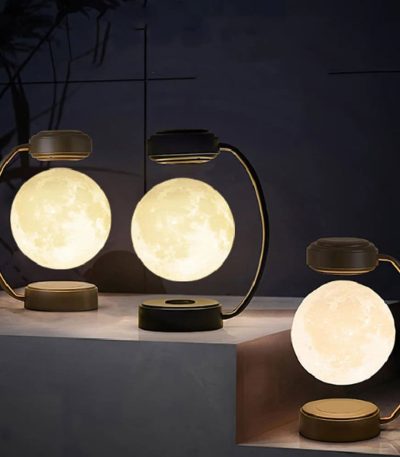 Três candeeiros lunares magnéticos com luz quente, base de metal dourado e design moderno, colocados sobre uma superfície de mármore escuro, com fundo escuro e plantas ao fundo.