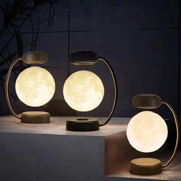 Três candeeiros lunares magnéticos com luz quente, base de metal dourado e design moderno, colocados sobre uma superfície de mármore escuro, com fundo escuro e plantas ao fundo.
