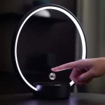 Lâmpada de plasma circular com anel de luz LED e esfera suspensa, manipulada por uma mão, em ambiente escuro.