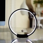 Candeeiro circular com luz LED branca, base preta e esfera metálica suspensa no centro, em ambiente de sala moderna.