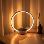 Candeeiro de mesa com estrutura circular de luz LED, base preta com botão de controlo, colocado numa mesa de madeira junto a uma jarra com palitos de incenso.