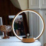 Candeeiro de mesa com estrutura circular de madeira e luz LED integrada, com base magnética e esfera de metal suspensa no centro.