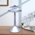 Candeeiro com design de disco voador em levitação, com luz azul, colocado numa mesa de madeira, com plantas e livros ao fundo.