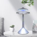 Candeeiro com design de ovni em levitação, com luz azul, colocado numa mesa branca ao lado de uma planta e um calendário.