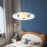 Candeeiro de teto com design de planeta, iluminação LED circular, com detalhes em dourado e branco, suspenso por cabos finos, em quarto com parede azul e cama de cabeceira de couro.