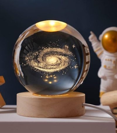 Globo de vidro transparente com representação de uma galáxia dourada e estrelas, colocado sobre uma base de madeira, com um astronauta de brinquedo ao fundo.