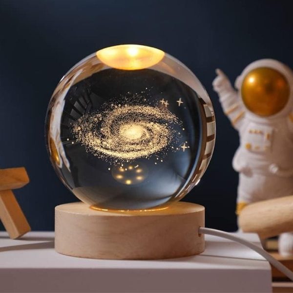 Globo de vidro transparente com representação de uma galáxia dourada e estrelas, colocado sobre uma base de madeira, com um astronauta de brinquedo ao fundo.