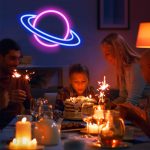 Família e amigos ao redor de uma mesa com bolo de aniversário, decorada com tema de espaço, incluindo uma iluminação de neon de um planeta com anéis.