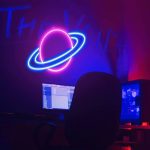 Luz de néon em forma de planeta com anel, em cores rosa e azul, iluminando um espaço escuro com um computador e uma cadeira de escritório.