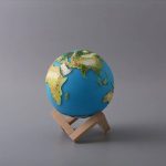 Mini globo terrestre com relevo detalhado, apoiado num suporte de madeira simples, sobre fundo cinzento.
