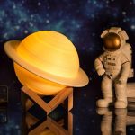 Luminária em forma de Saturno com anéis iluminados, acompanhada de um astronauta de brinquedo com traje espacial detalhado, fundo de estrelas.