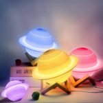 Três lâmpadas decorativas em forma de Saturno com diferentes cores de luz, uma amarela, uma azul e uma rosa, em uma superfície com acessórios decorativos.
