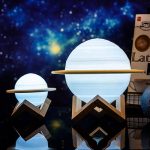 Lâmpada decorativa em forma de planeta Saturno com anel, iluminada com luz azul, colocada sobre suporte de madeira, fundo de espaço estrelado.