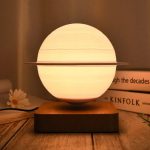 Candeeiro com forma de Saturno, com anel metálico ao redor, iluminado, sobre base de madeira, ambiente acolhedor.