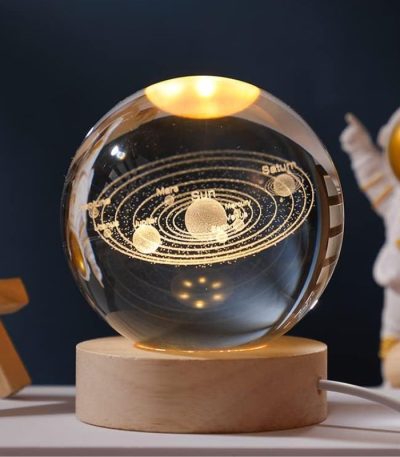 Globo de vidro transparente com representação dourada do sistema solar, incluindo planetas e órbitas, apoiado numa base de madeira clara.