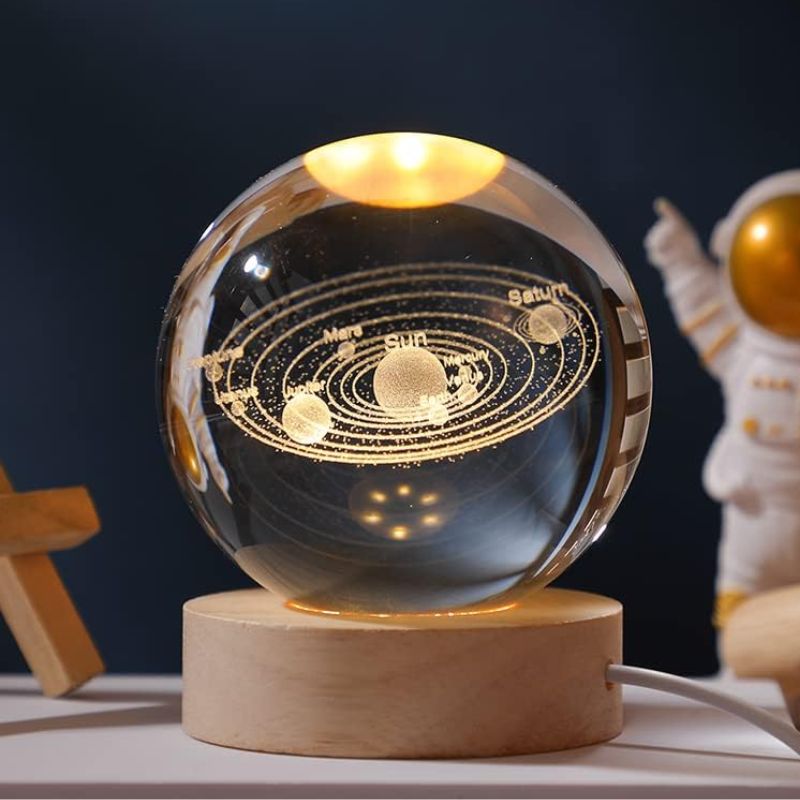 Globo de vidro transparente com representação dourada do sistema solar, incluindo planetas e órbitas, apoiado numa base de madeira clara.