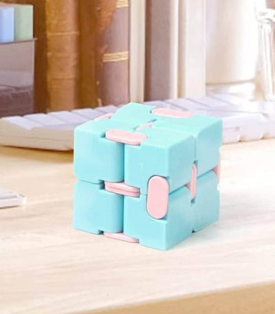 Cubo fidget infinito com blocos azuis e detalhes rosa, mostrado numa mesa de madeira clara, com fundo de estantes e objetos de escritório.