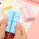 Cubo fidget infinito em cores pastel azul, rosa e amarelo, com partes articuladas que se movem facilmente, mostrado na mão e em uma mesa com outros cubos semelhantes.