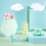 Vários cubos fidget em cores pastel, alguns empilhados, outros dispostos em diferentes posições, com fundo de nuvens de papel e elementos geométricos.