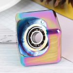Cubo fidget com acabamento iridescente, detalhes do mecanismo interno visíveis, em fundo branco com objetos de escritório ao lado.