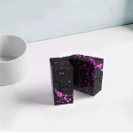 Cubo infinito de aço preto com manchas de tinta rosa, exibido numa superfície branca ao lado de uma chávena branca.