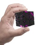 Pequeno cubo de aço preto com pintura rosa a spray, mostrado na mão de uma pessoa, com detalhes de pintura dispersa na superfície.