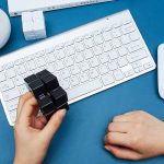 Mão segurando cubo infinito anti-stress preto com várias faces, perto de teclado branco e acessórios de escritório.