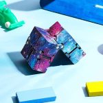 Cubo Infinito Galáxia com padrão de galáxia, detalhes em roxo, azul e rosa, com várias faces visíveis, colocado numa superfície azul clara com outros brinquedos ao redor.
