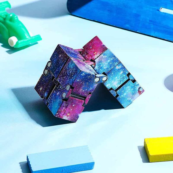 Cubo Infinito Galáxia com padrão de galáxia, detalhes em roxo, azul e rosa, com várias faces visíveis, colocado numa superfície azul clara com outros brinquedos ao redor.