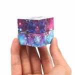 Pequeno cubo com padrão de galáxia, com cores vibrantes de roxo, azul e rosa, sendo segurado por uma mão, mostrando detalhes do padrão espacial e do mecanismo de encaixe.