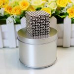 Cubo de bolas magnéticas prateadas empilhadas formando um cubo tridimensional, com fundo de flores amarelas e brancas em uma caixa de madeira branca.