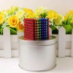 Cubo magnético com várias cores, detalhes de esferas metálicas, colocado sobre uma caixa metálica prateada, com flores amarelas ao fundo.
